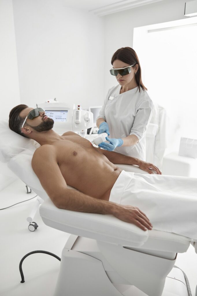 Young master with goggles conducts laser epilation to bearded man client lying on couch in clinic